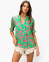 A woman modeling a bright green and pink abstract patterned short-sleeve blouse with a split neckline, paired with light beige frayed denim shorts, against a plain white background.