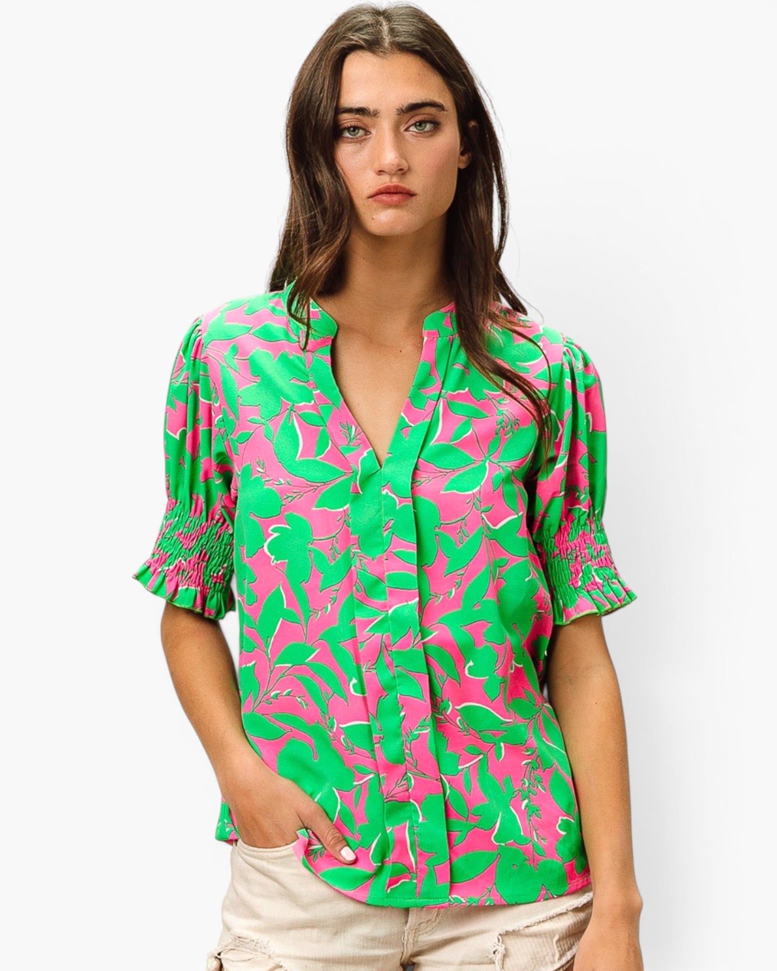 A woman modeling a short-sleeve button-up blouse with a vibrant green and pink abstract pattern, paired with light beige pants, against a plain light gray background.