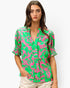 A woman modeling a short-sleeve button-up blouse with a vibrant green and pink abstract pattern, paired with light beige pants, against a plain light gray background.