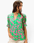A woman wearing a short-sleeve button-up blouse with a bold pink and green abstract pattern, paired with light beige pants, against a plain white background.