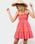 A woman wearing a coral pink tiered mini dress with ruffled straps and a wide-brimmed straw hat, posed against a light gray studio background.