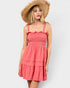 A woman wearing a coral pink smocked tiered dress with ruffled straps and a wide-brimmed straw hat, photographed against a light gray studio background.