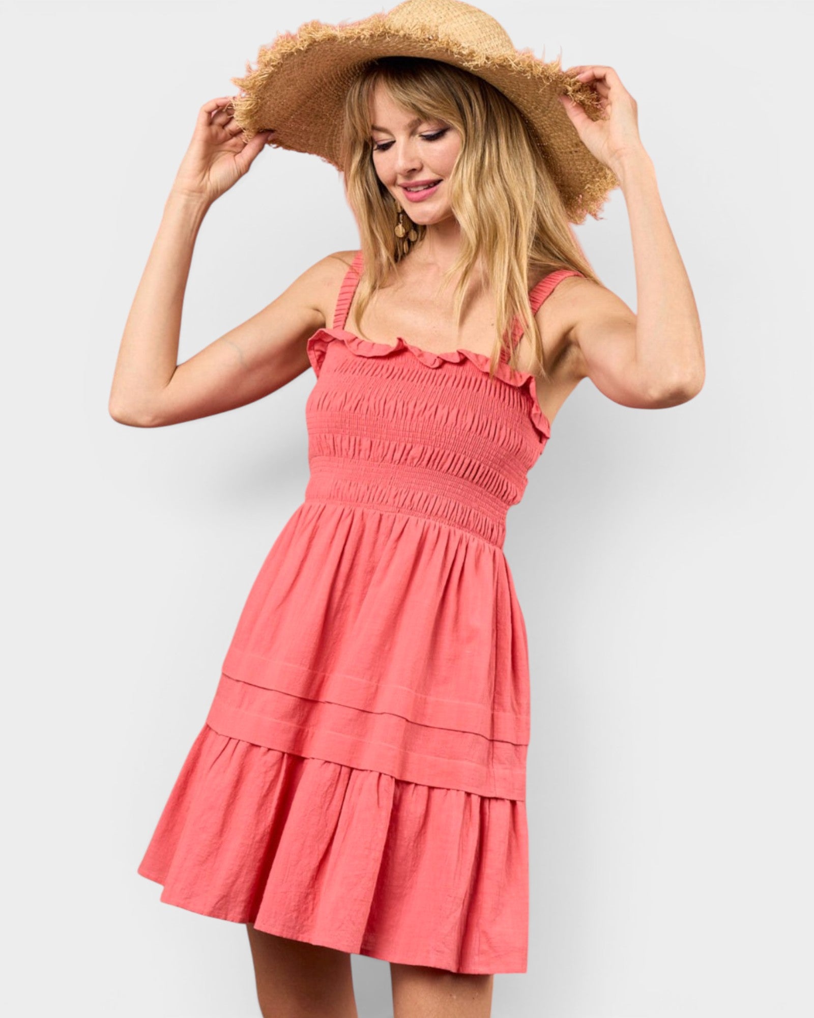 A woman wearing a coral pink tiered smocked dress and a wide-brimmed straw hat, posing against a light gray studio background.