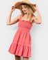 A woman wearing a coral pink tiered smocked dress and a wide-brimmed straw hat, posing against a light gray studio background.