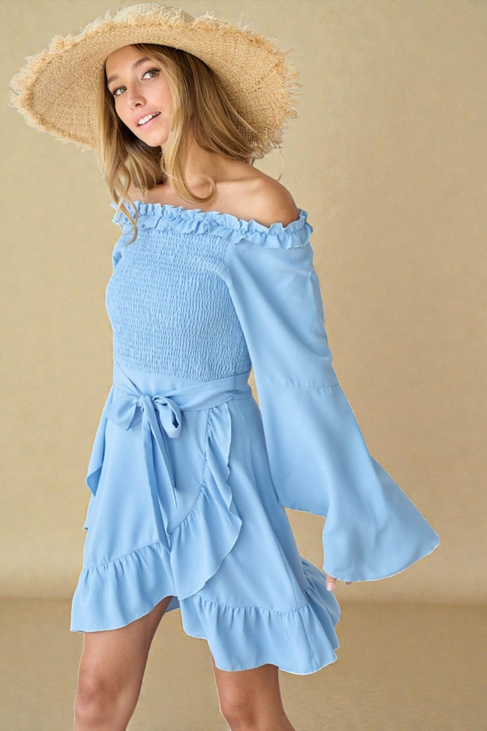 Woman wearing a light blue off-shoulder dress with ruffled details and a straw hat against a beige background.