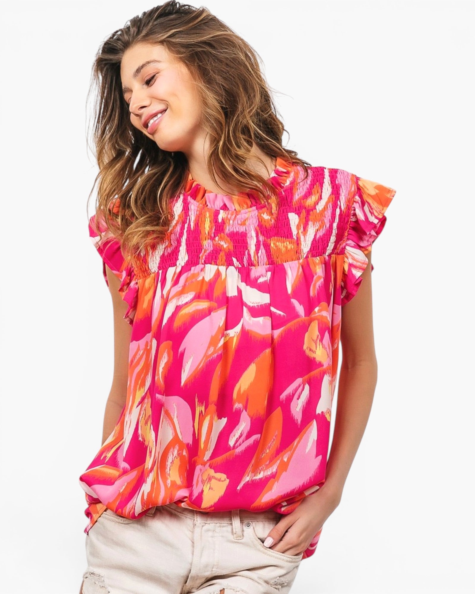 A woman wearing a bright pink short-sleeve blouse with an abstract orange, pink, and white floral print, paired with light beige pants, against a plain white background.