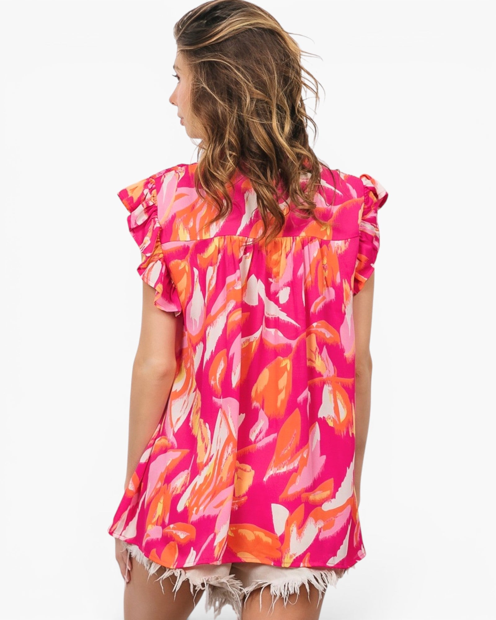 A woman shown from the back wearing a bright pink short-sleeve top with an abstract orange, yellow, and white floral print, paired with frayed denim shorts, against a plain white background.