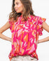 A woman wearing a bright pink and orange abstract print blouse with ruffled sleeves, paired with light beige jeans, against a plain white background.