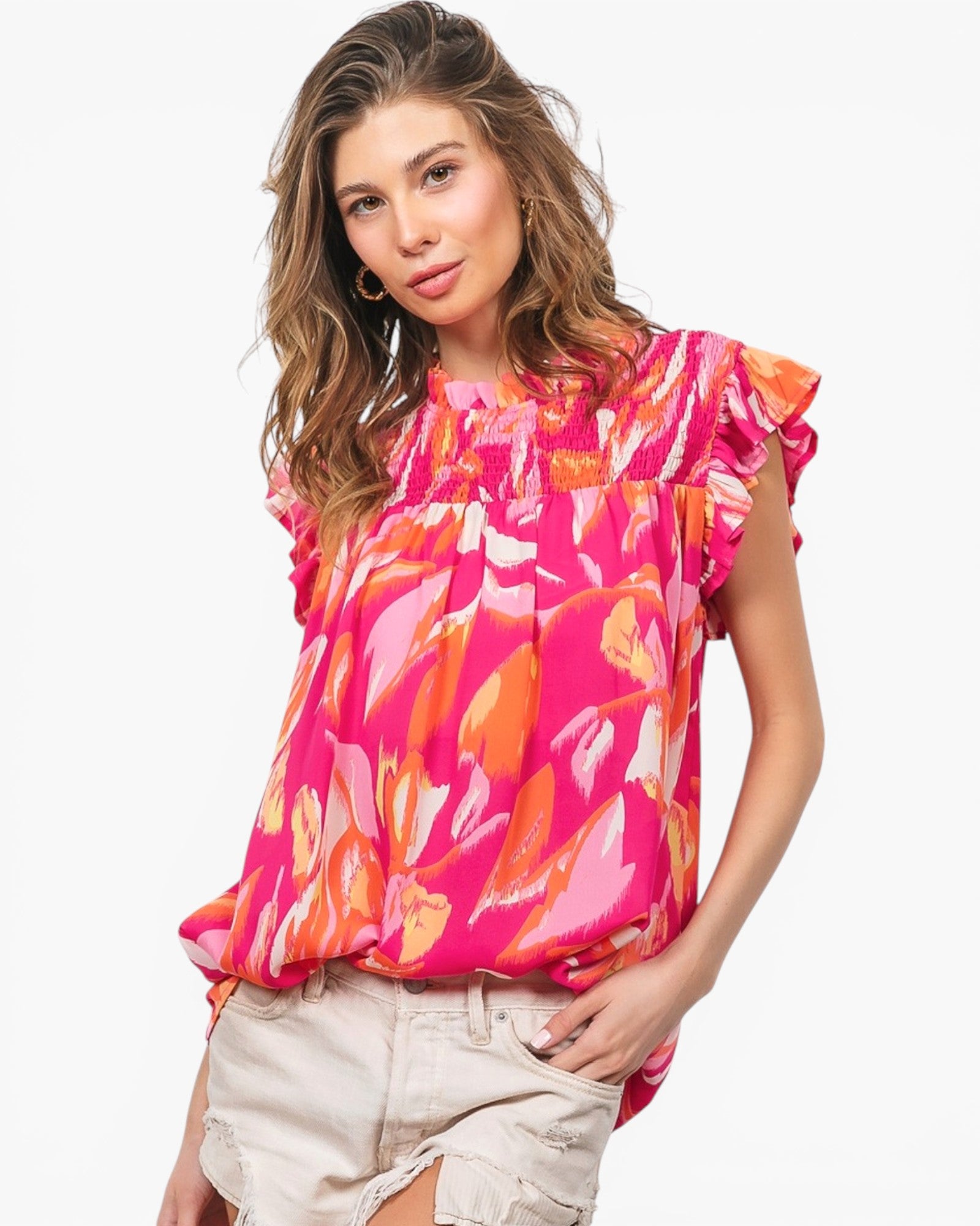 A woman wearing a bright pink and orange abstract print ruffle-sleeve blouse with light beige shorts, photographed against a plain white studio background.