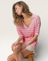 Woman wearing a pink striped shirt sitting on a wooden stool against a plain background