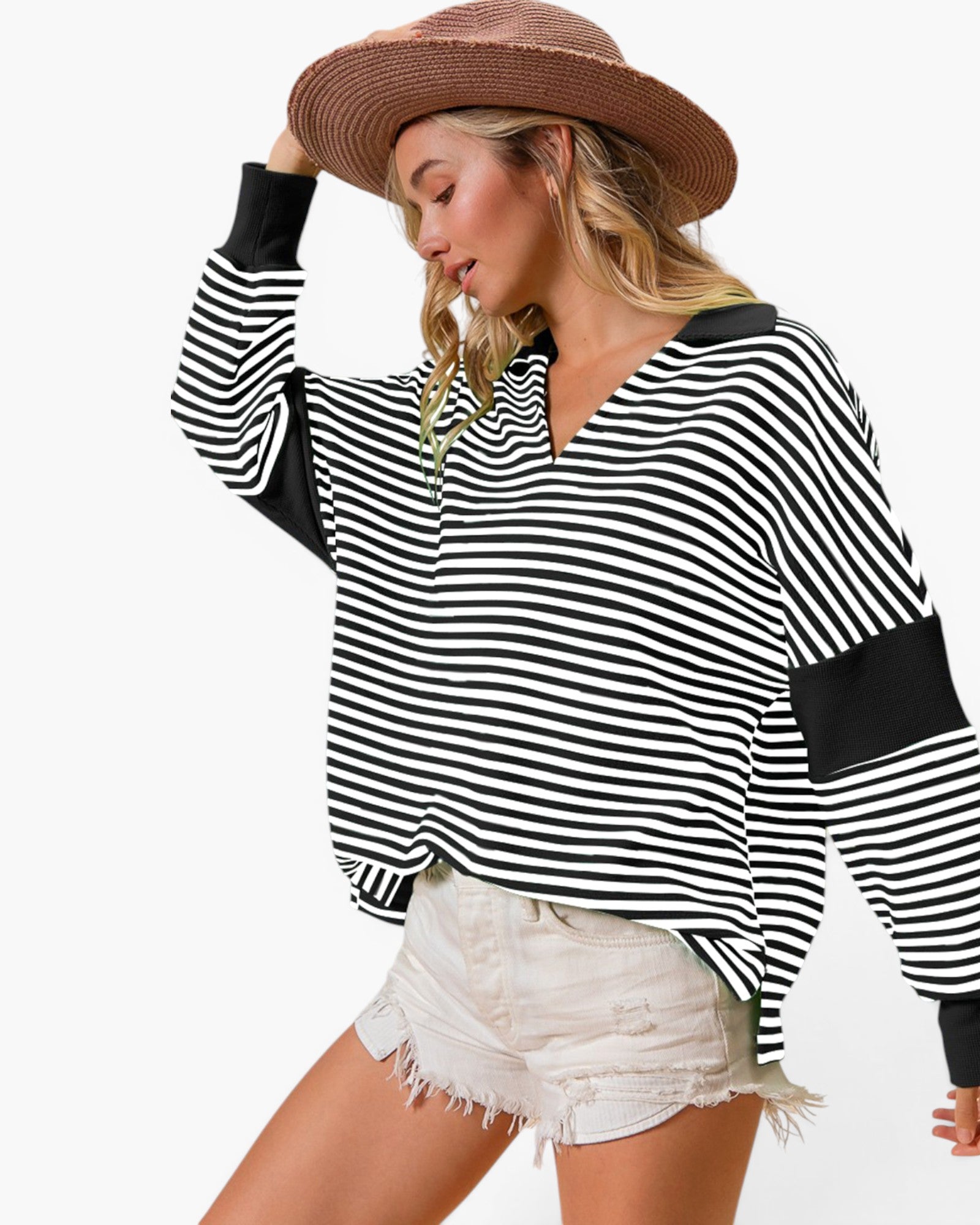 A woman wearing a black and white horizontally striped long-sleeve top with solid black contrast panels on the shoulders and cuffs, paired with distressed white denim shorts and a brown straw hat, against a plain white background.