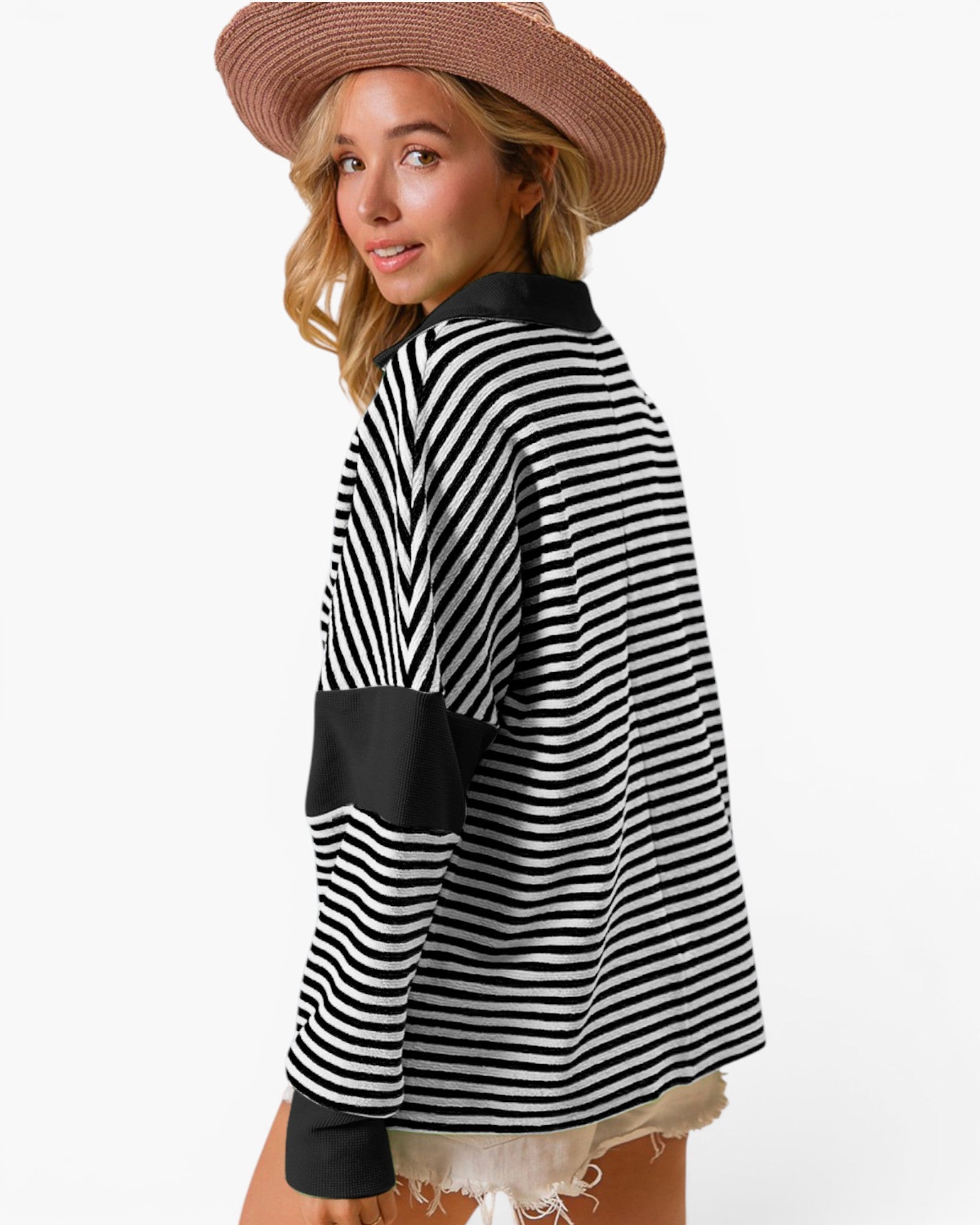 A woman wearing a black and white horizontally striped long-sleeve top with solid black color-blocked panels on the collar, cuffs, and sleeve, paired with a brown wide-brim hat and light denim shorts, shown from a three-quarters back view against a white studio background.
