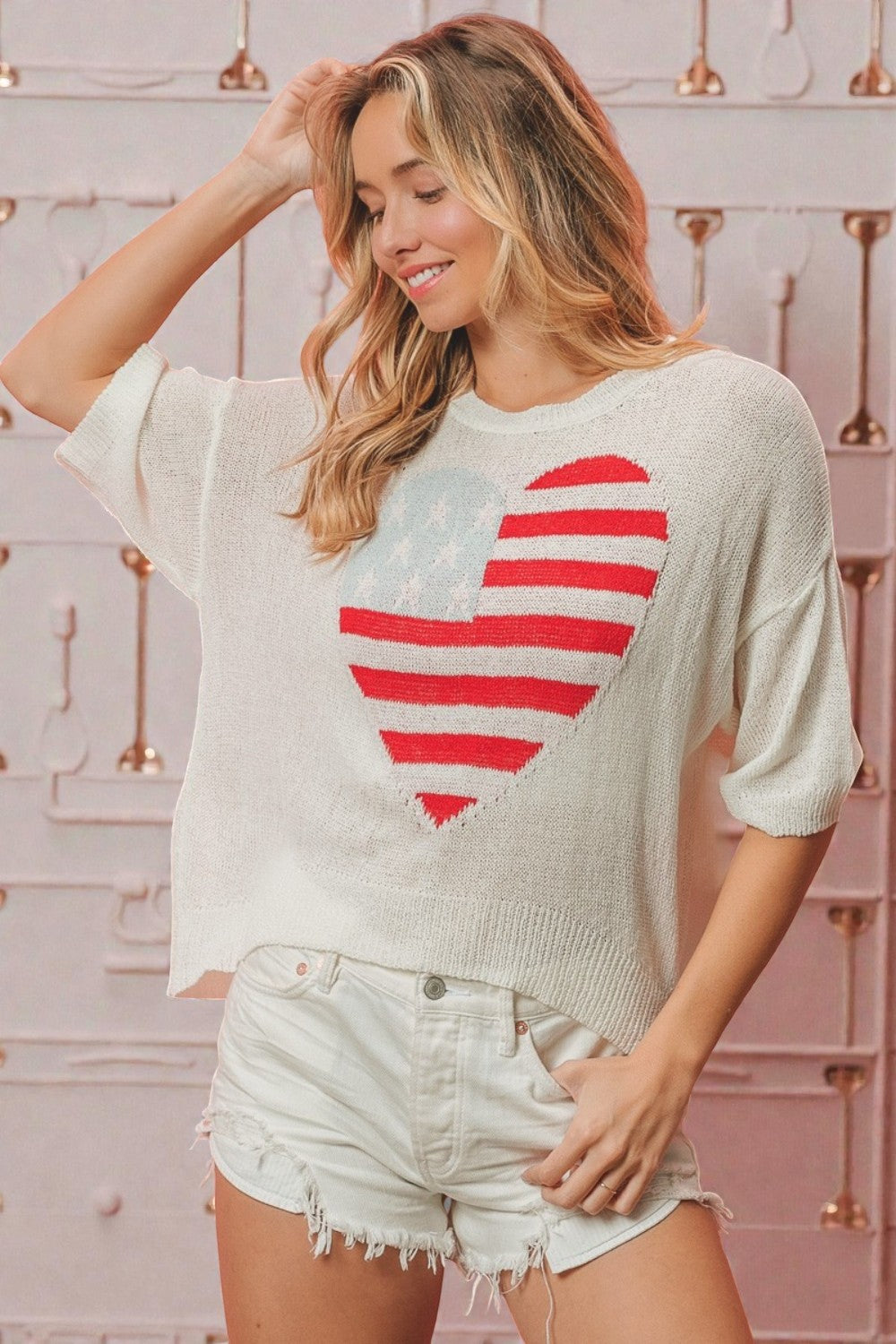 Woman wearing a white sweater with a red heart design and denim shorts against a decorative wall.