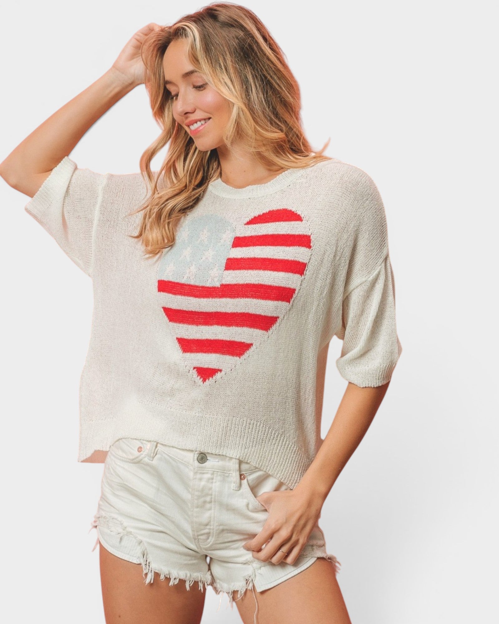 A woman wearing a cream-colored knit sweater with a distressed red and white heart graphic on the front, paired with light denim shorts, against a light gray studio background.