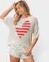 A woman wearing a cream-colored knit sweater with a distressed red and white heart graphic on the front, paired with light denim shorts, against a light gray studio background.
