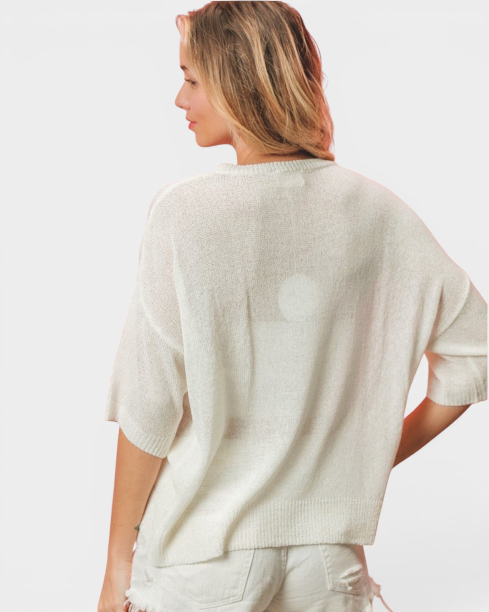A woman shown from the back wearing a loose, short-sleeve knit top in a light cream or off-white color, paired with light-colored pants, against a plain light gray studio background.