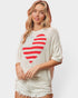 A woman wearing a cream-colored knit sweater with a large red and white striped heart graphic on the front, paired with distressed denim shorts, against a light gray studio background.