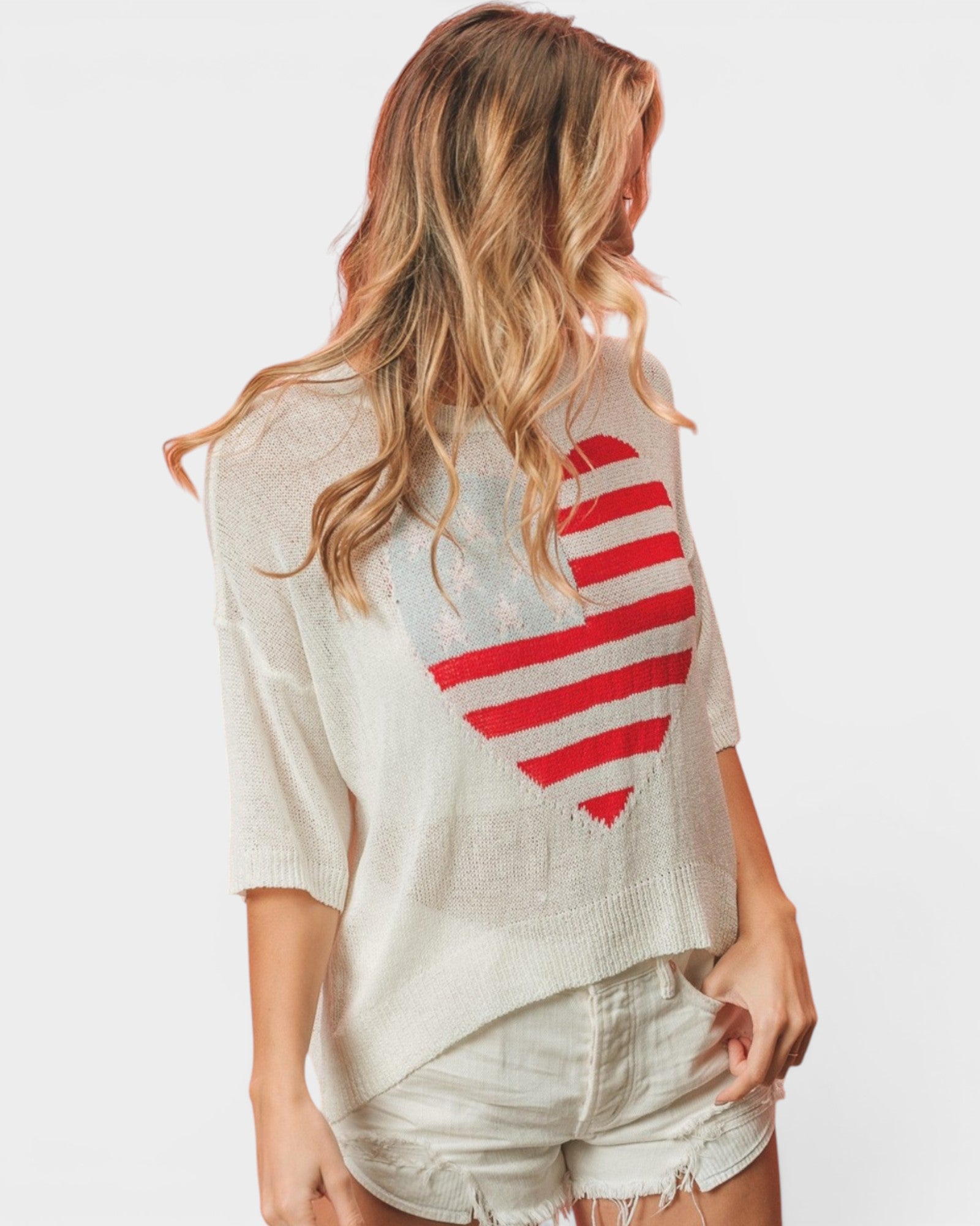 A woman shown from the back wearing a distressed off-white knit sweater with a large red and white striped heart graphic on the back, paired with light denim shorts, against a plain light gray background.