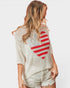 A woman shown from the back wearing a distressed off-white knit sweater with a large red and white striped heart graphic on the back, paired with light denim shorts, against a plain light gray background.