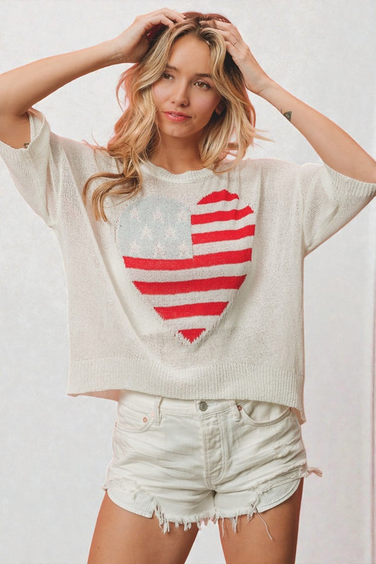 Woman wearing a white sweater with a red heart design and denim shorts on a plain background