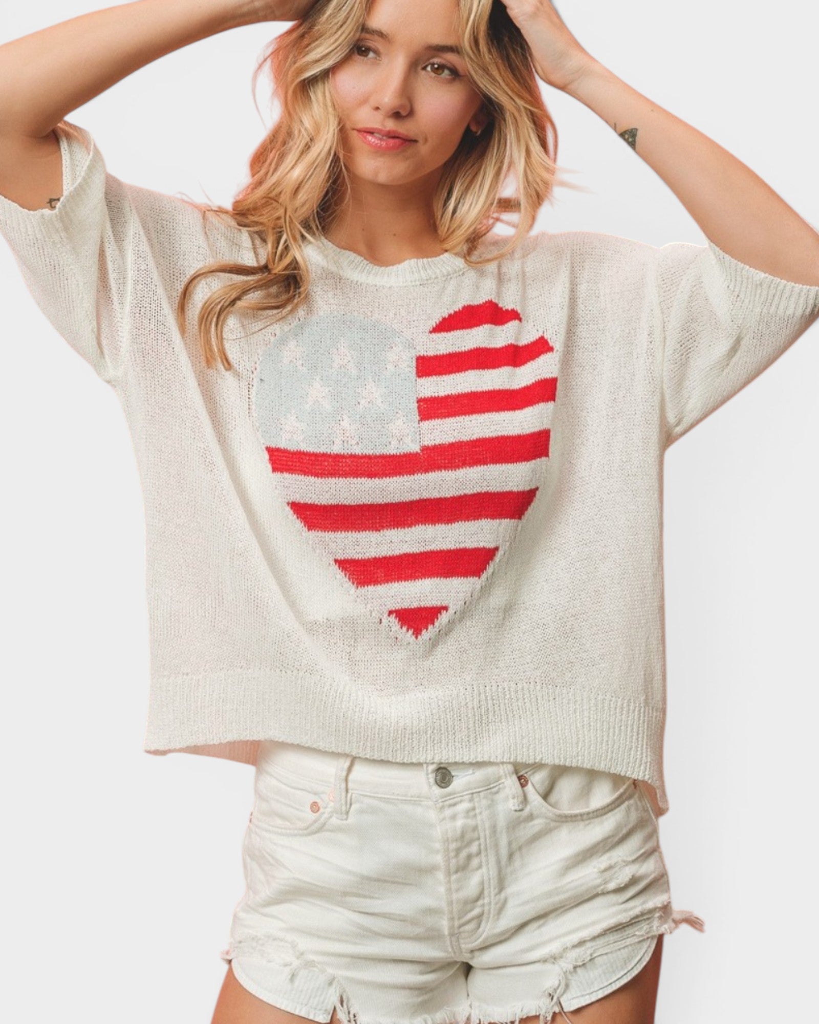 A woman wearing a cream-colored knit sweater with a large heart-shaped American flag graphic on the front, paired with light-wash denim shorts, against a plain light gray background.