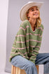 Woman wearing a green and pink striped sweater with a white cowboy hat and blue jeans, sitting on a wooden stool.