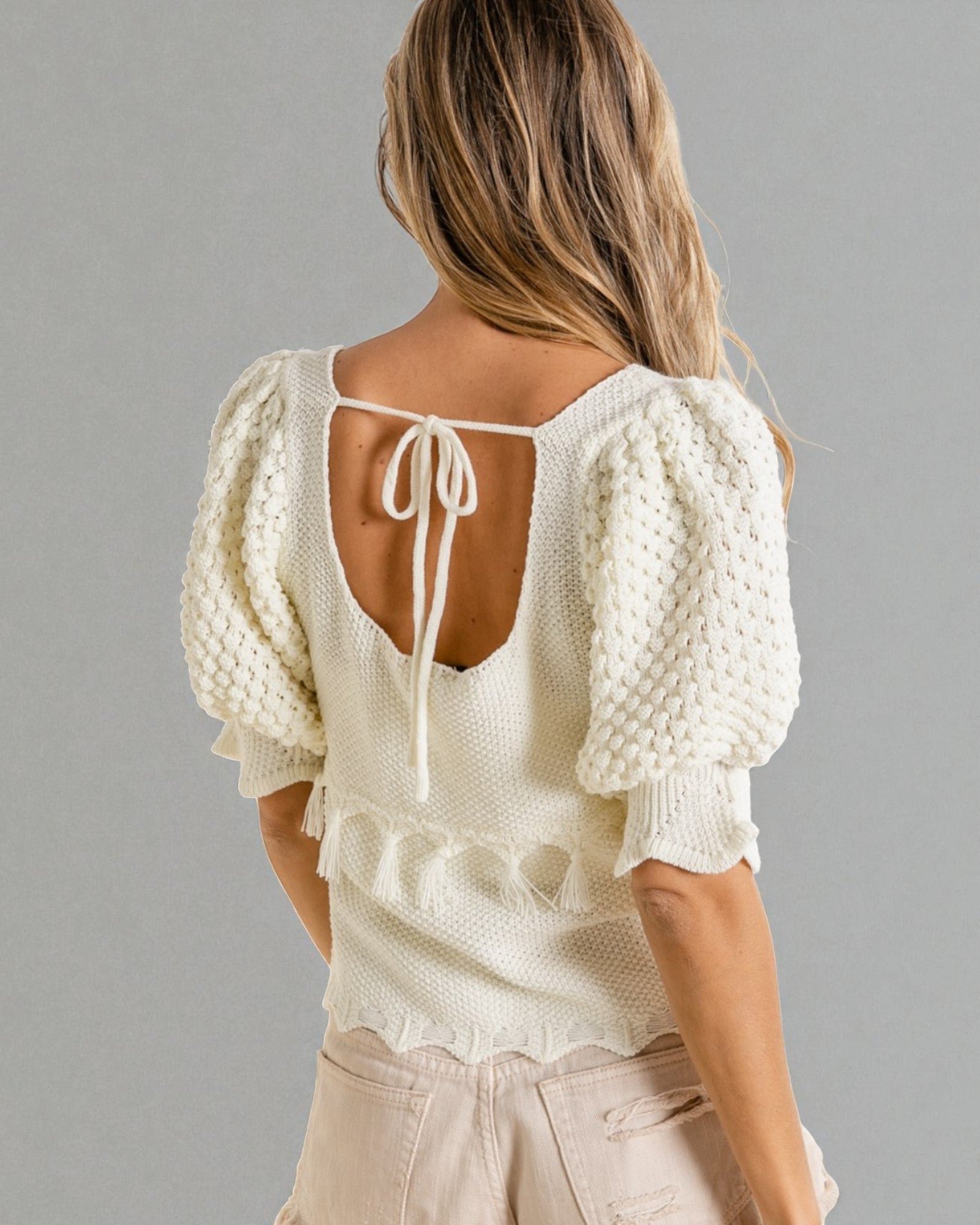 Woman wearing a white textured blouse with puff sleeves and a open back on a gray background