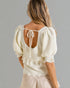 Woman wearing a white textured blouse with puff sleeves and a open back on a gray background