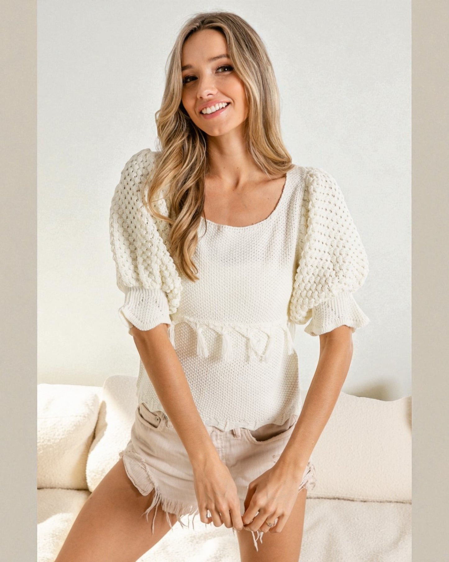 Woman wearing a white textured top and denim shorts sitting on a couch.