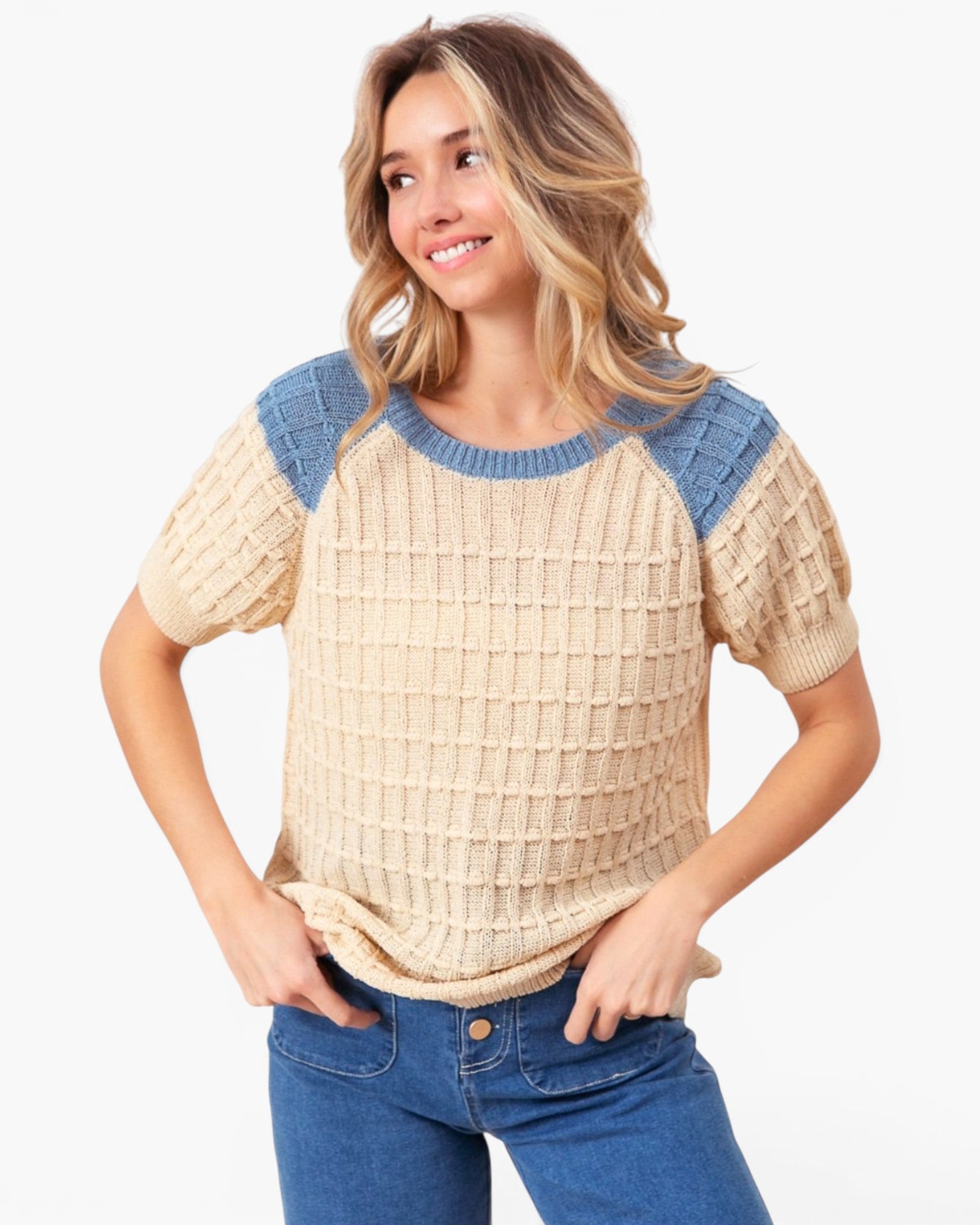A woman wearing a beige and blue short-sleeve knit sweater with textured waffle patterns, paired with blue jeans, against a plain white background.