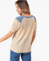 A woman shown from the back wearing a short-sleeve knit top with a beige body, textured diamond-pattern sleeves, and blue ribbed shoulder yokes, paired with blue jeans against a light gray studio background.