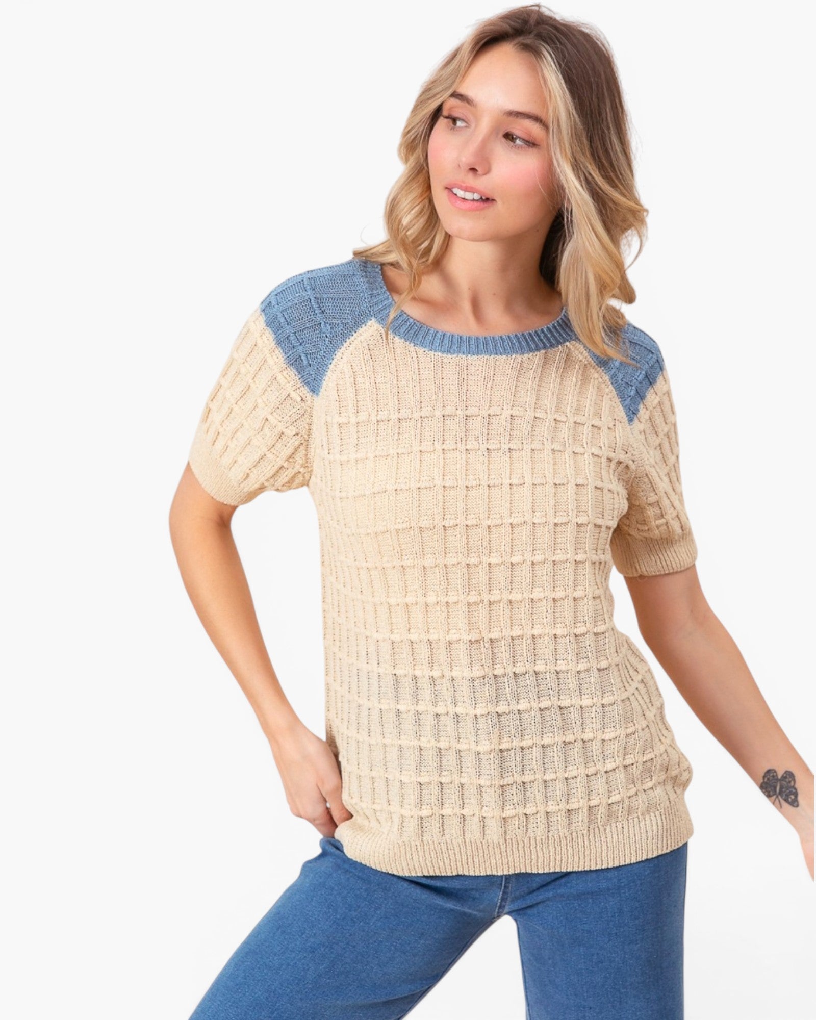 A woman wearing a short-sleeve knit top with a beige waffle texture and blue raglan shoulder panels, paired with blue jeans, against a plain white background.