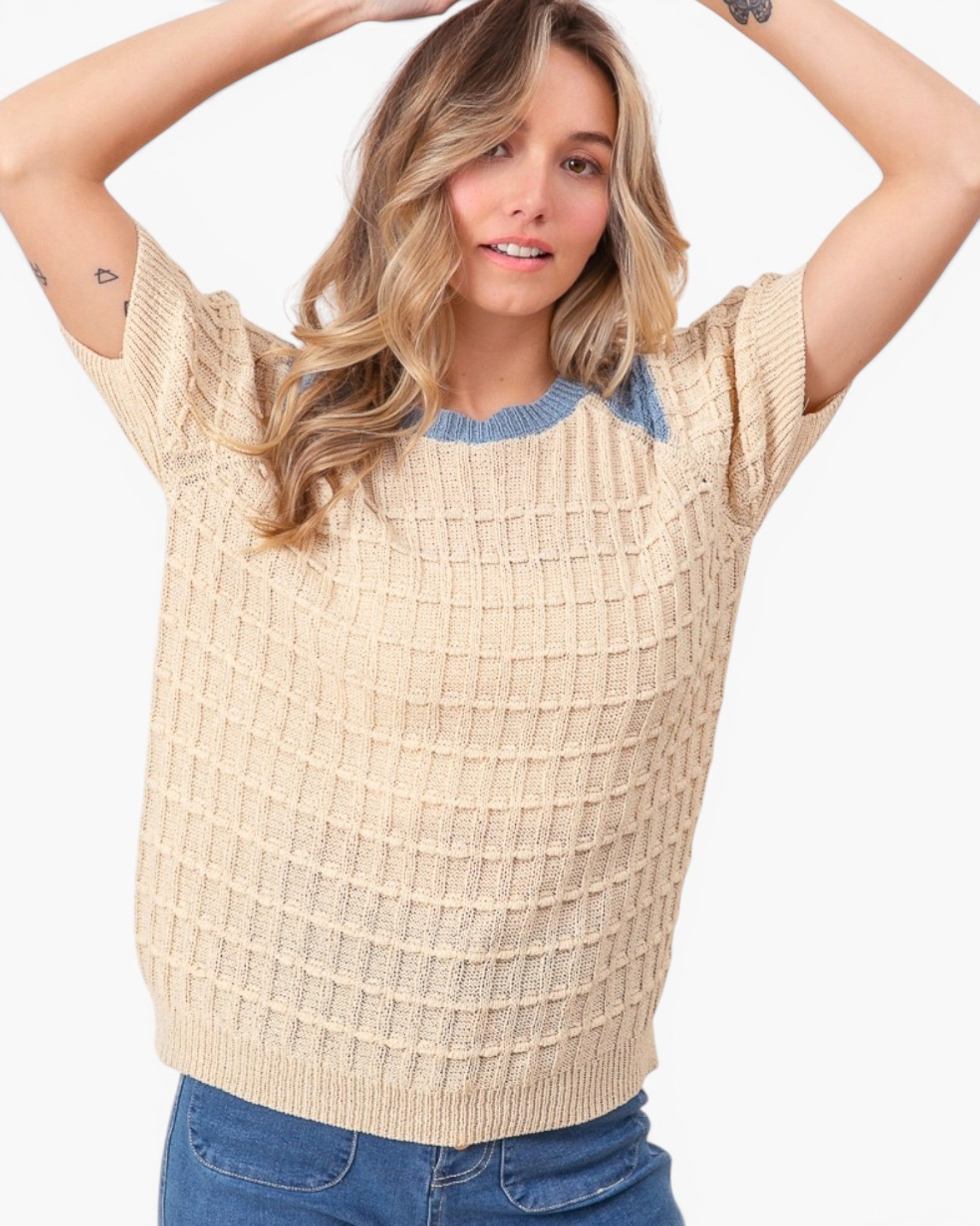 A woman wearing a beige short-sleeve knit sweater with a blue crew neck collar, paired with blue jeans, against a plain light gray background.
