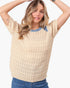 A woman wearing a beige short-sleeve knit sweater with a blue crew neck collar, paired with blue jeans, against a plain light gray background.