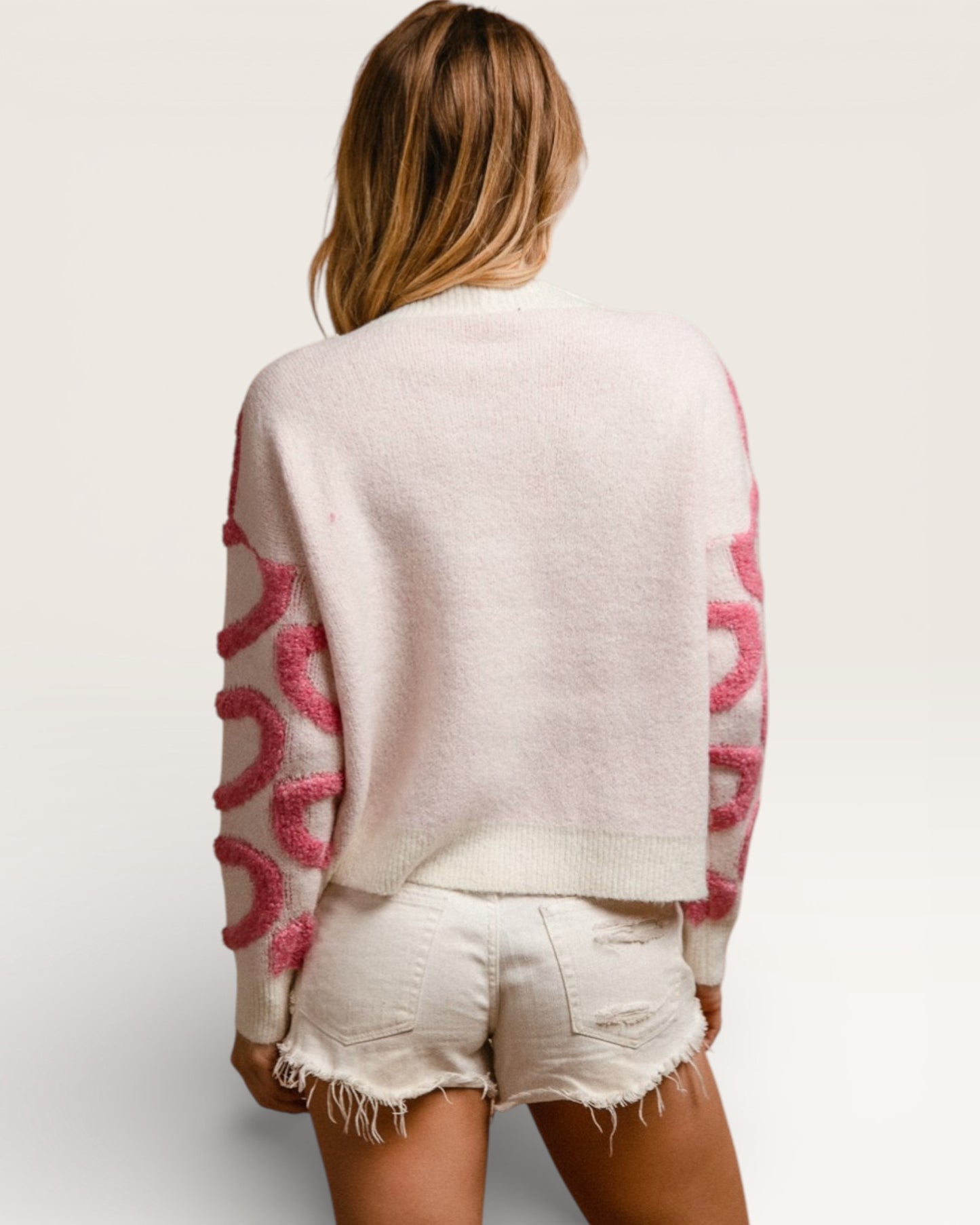 Person wearing a light-colored sweater with pink patterns and white shorts on a plain background