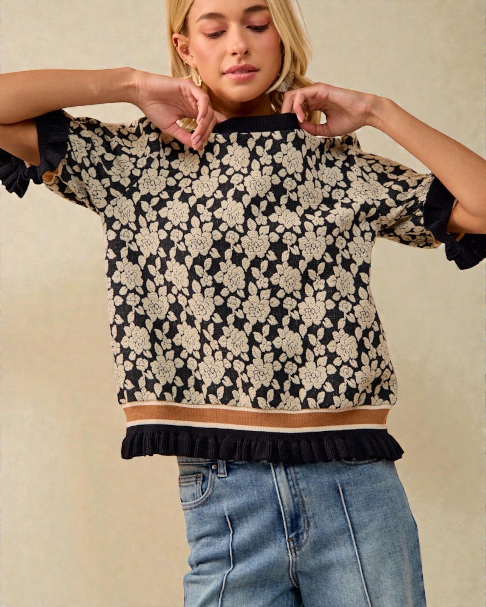 Woman wearing a black floral blouse with ruffled sleeves and hem, standing against a beige background.