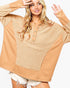 A woman wearing a tan and beige patchwork long-sleeve henley top with a textured knit center panel and smooth side panels, paired with white frayed denim shorts, against a plain white background.