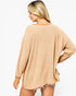 A woman shown from the back wearing a loose, oversized knit sweater in a warm beige or tan color, photographed against a plain light gray studio background.
