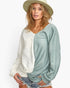 A woman wearing a color-block knit sweater with one half in textured cream and the other half in dusty sage green, paired with denim shorts and a wide-brimmed beige hat against a white studio background.