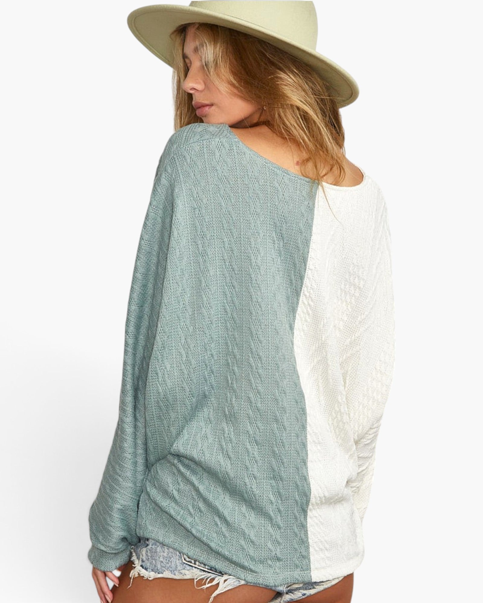 A woman shown from the back wearing a color-block knit top with a sage green left side and an off-white right side, paired with a light beige wide-brim hat and denim shorts against a plain white background.