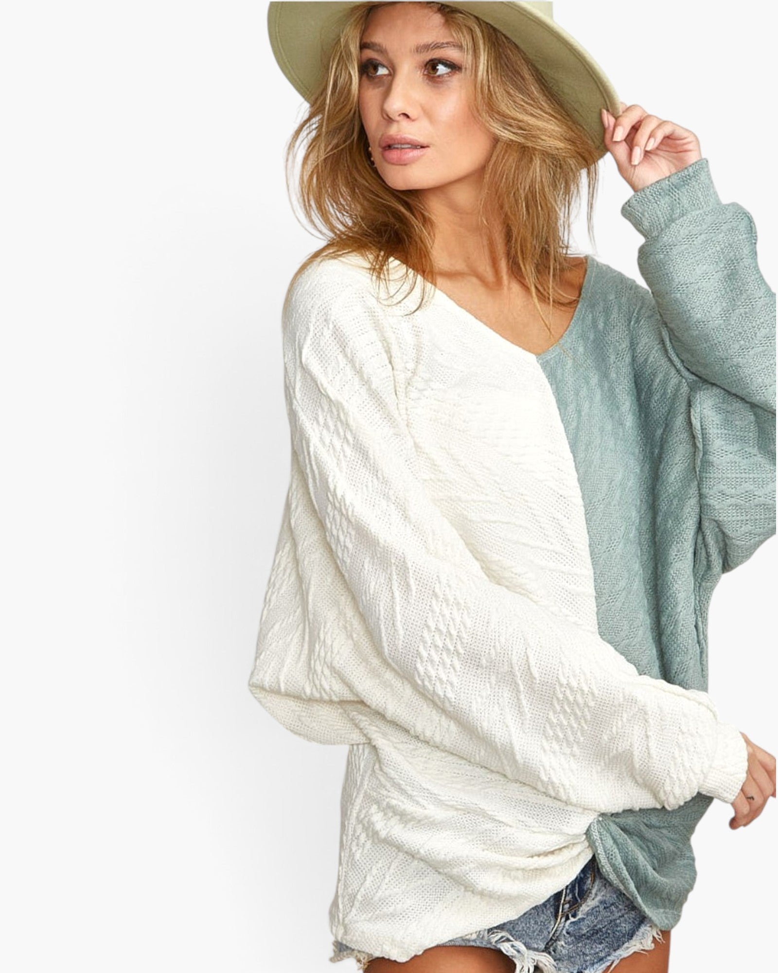 A woman wearing a color-block knit sweater with a textured cream body and a sage green sleeve, paired with denim shorts and a wide-brimmed hat against a plain white background.
