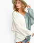 A woman wearing a color-block knit sweater with a textured cream body and a sage green sleeve, paired with denim shorts and a wide-brimmed hat against a plain white background.