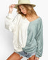 A woman wearing a split-color long-sleeve knit top with one half in textured cream and the other half in dusty sage green, styled with denim shorts and a beige wide-brim hat against a white studio background.