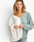 A woman modeling a long-sleeve V-neck knit top with a color-block design in cream and sage green, paired with denim shorts against a light gray studio background.