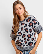 A woman wearing a short-sleeve knit sweater with a leopard print pattern in black, brown, and beige on a light gray base, featuring striped ribbed cuffs and hem.