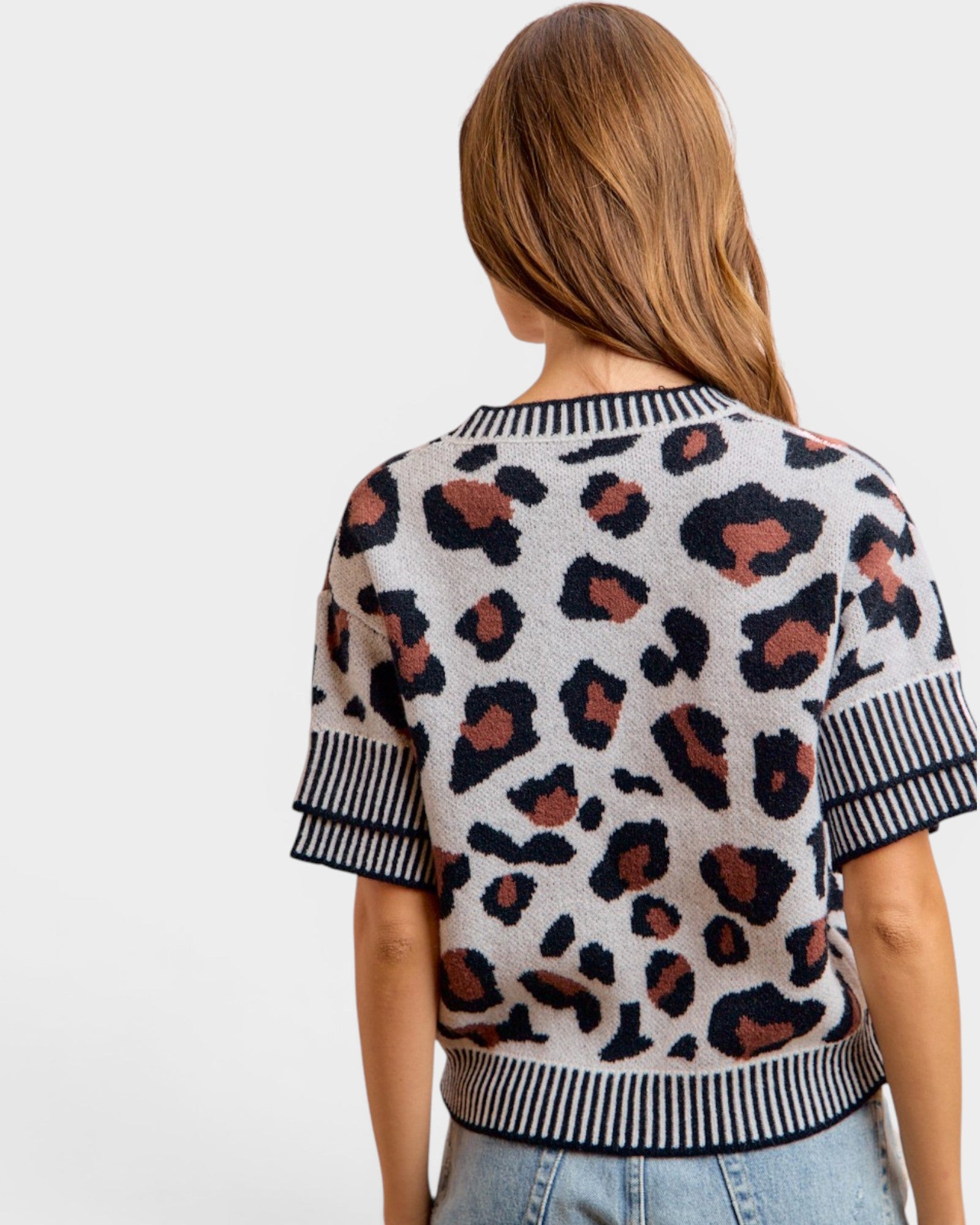 A woman shown from the back wearing a short-sleeve knit top with a leopard print pattern on a light beige background with black and brown spots, featuring striped ribbed cuffs and hem.