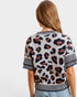 A woman shown from the back wearing a short-sleeve knit top with a leopard print pattern on a light beige background with black and brown spots, featuring striped ribbed cuffs and hem.