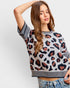 A woman wearing a short-sleeve knit sweater with a leopard print pattern in brown, black, and beige, featuring striped ribbed cuffs and hem, paired with blue jeans against a light gray studio background.