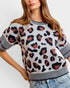 A woman wearing a short-sleeve knit sweater with a leopard print pattern in black, navy, and terracotta on a light gray base, paired with blue jeans against a plain light gray background.