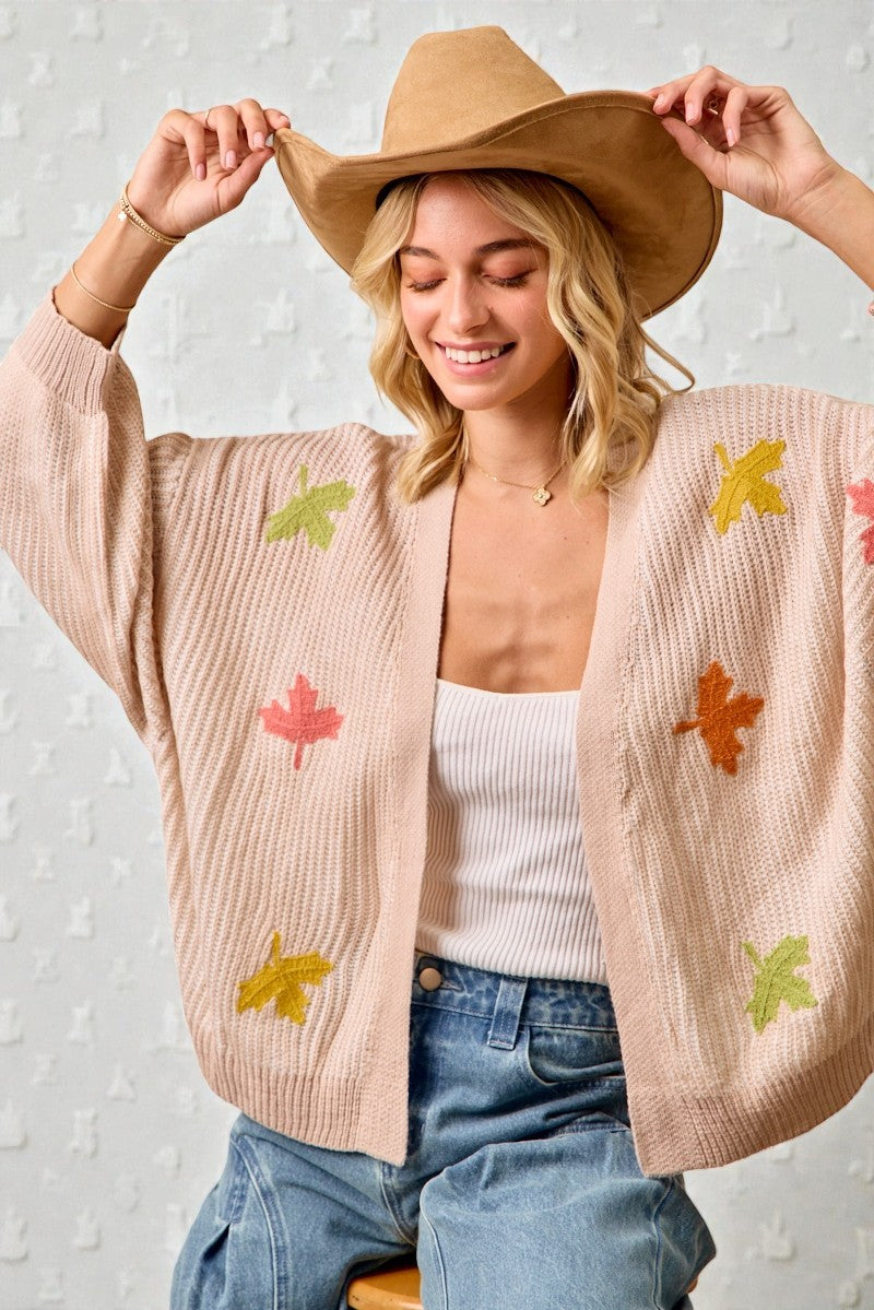 Woman wearing a beige cardigan with leaf patterns, white top, and blue jeans against a light background